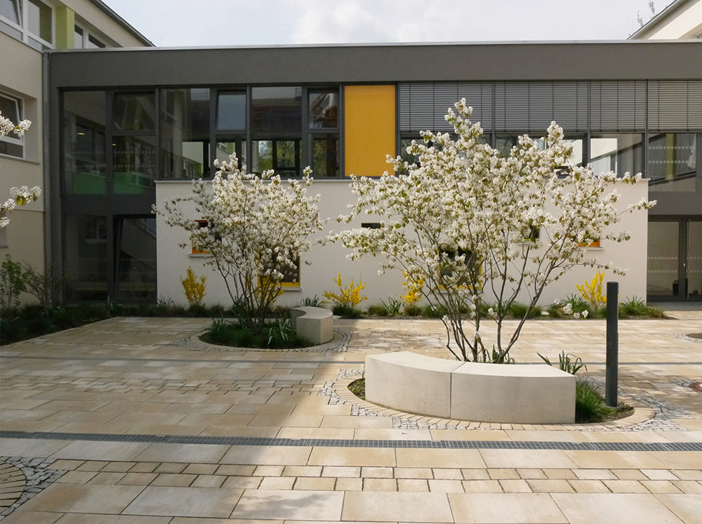 Ansicht der Außenanlagen des Dresdner Kindergartens "Bunte Kinderwelt" mit blühender Bepflanzung, Sitzmauern unter Kupferfelsenbirnen und einer Platzgestaltung mit unterschiedlich formatigen Natursteinplatten