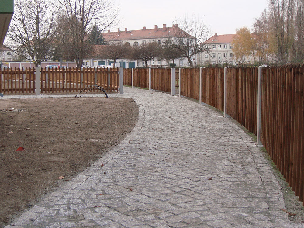 Erschließungsweg aus Natursteinpflaster, der durch eine mit Holzzäunen abgetrennte Gartenanlage führt